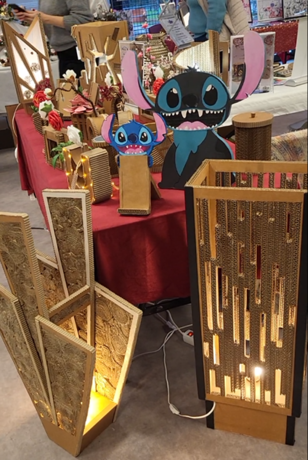 Stand de marché artisanal présentant des lampes décoratives en carton éclairées et des figurines colorées de Stitch, disposées sur une table recouverte d’une nappe rouge, avec des visiteurs en arrière-plan